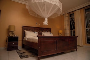 A bedroom with a wooden bed, bedside lamps, a nightstand, and a large window with curtains. - Pearl On The Nile Hotel Jinja (Jinja)