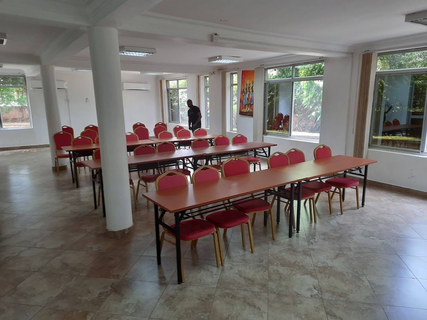 A spacious room with multiple tables and chairs, large windows, and a person standing in the background.