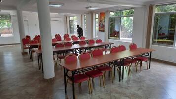 A spacious room with multiple tables and chairs, large windows, and a person standing in the background.