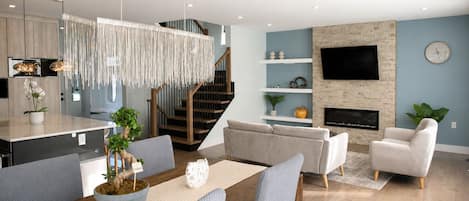 A modern living room with a sofa, armchairs, a dining table with chairs, a kitchen area, and a staircase.