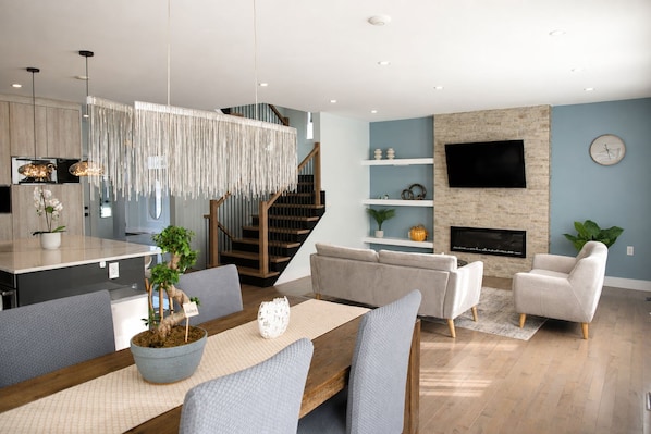 A modern living room with a sofa, armchairs, a dining table with chairs, a kitchen area, and a staircase.