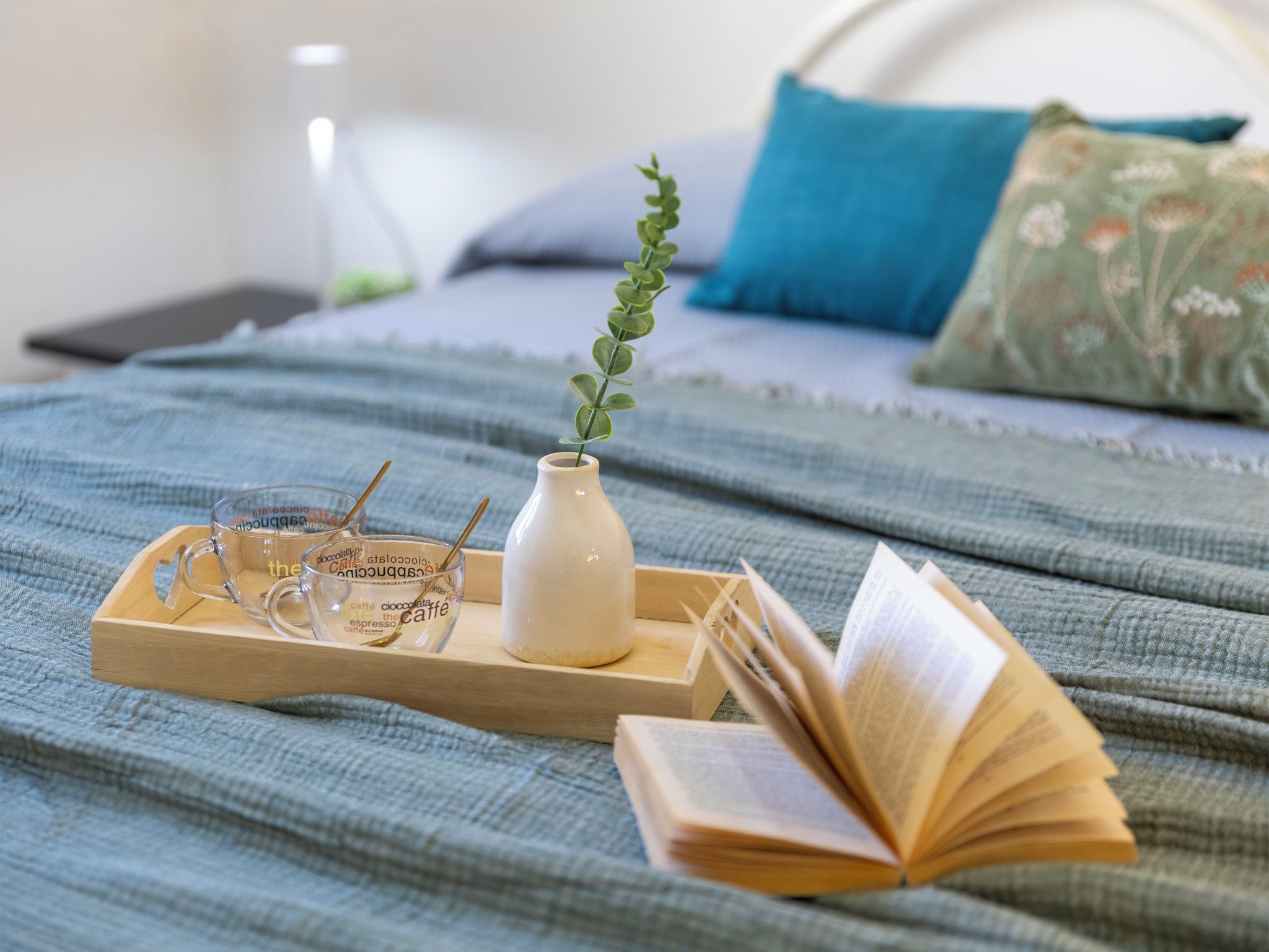 A bed with a blue blanket, a wooden tray with a glass cup and a white vase, and an open book.