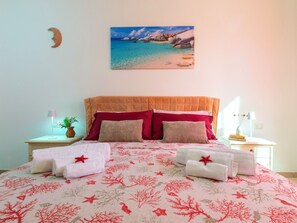 A bedroom with a bed, pillows, a nightstand, a lamp, a vase with flowers, and a painting of a beach scene. - Il Giardino di Sabrina by Interhome (Valledoria)