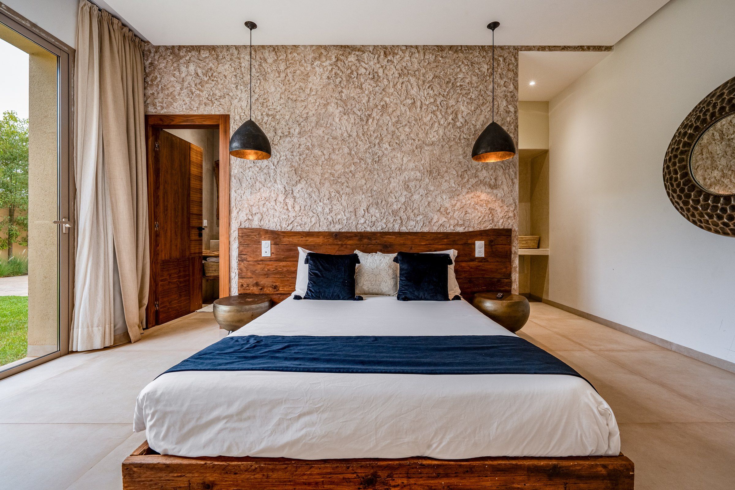 Une chambre avec un grand lit, une tête de lit en bois, deux lampes suspendues et une vue sur l’extérieur.