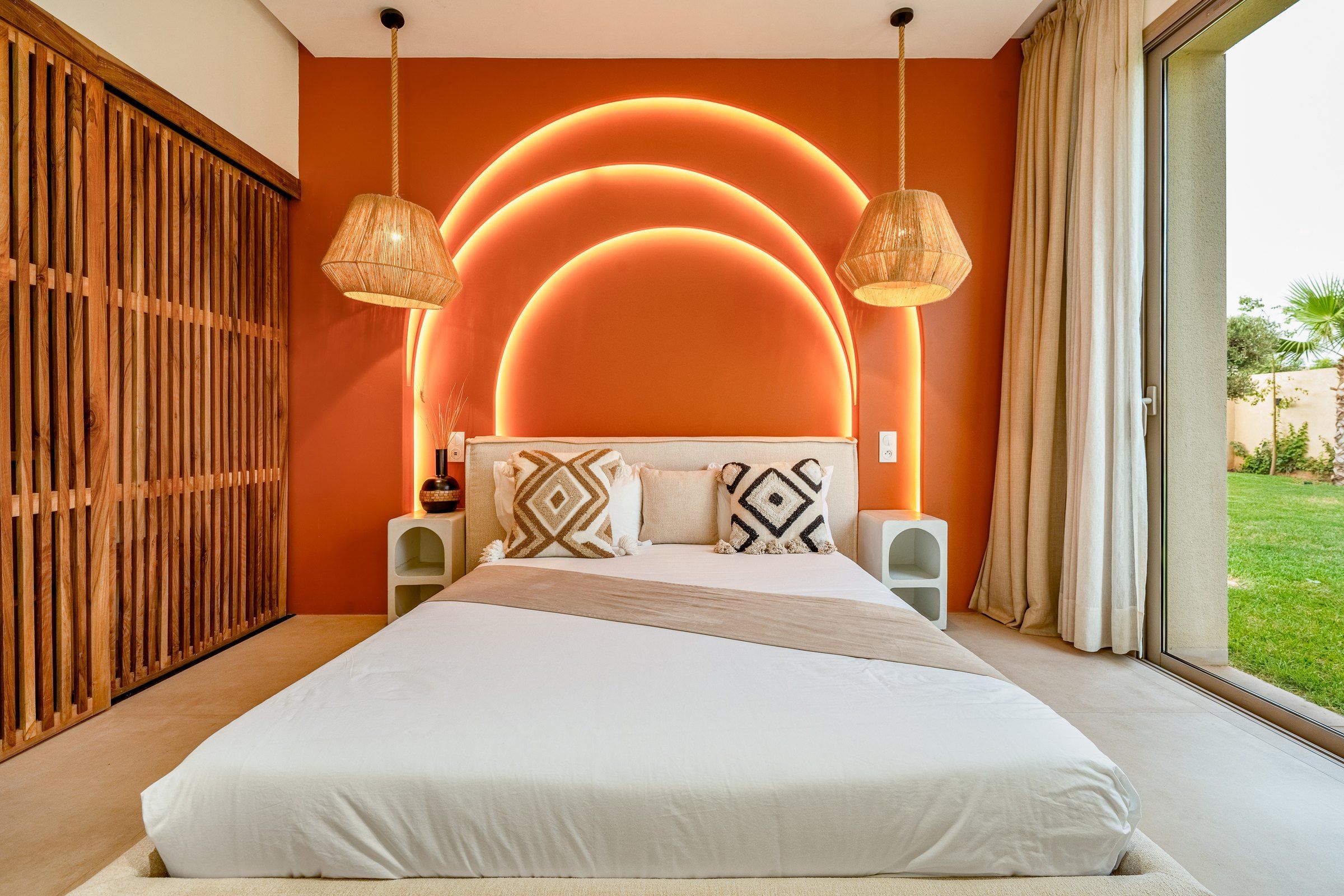 A modern bedroom with a large bed, two bedside tables, and a view of a garden through a sliding door.