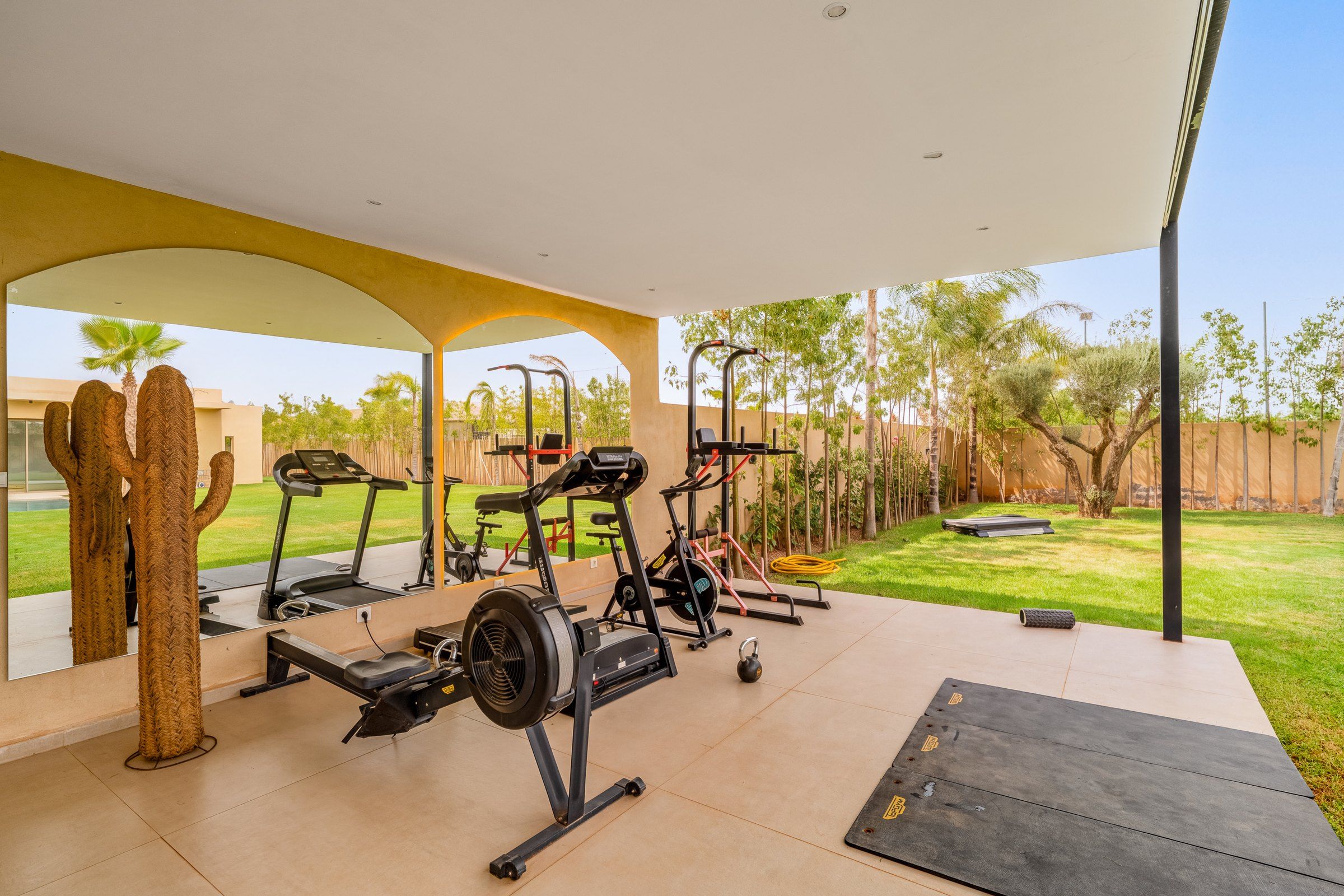 An outdoor gym area with exercise equipment, a covered walkway, and a view of a landscaped garden.