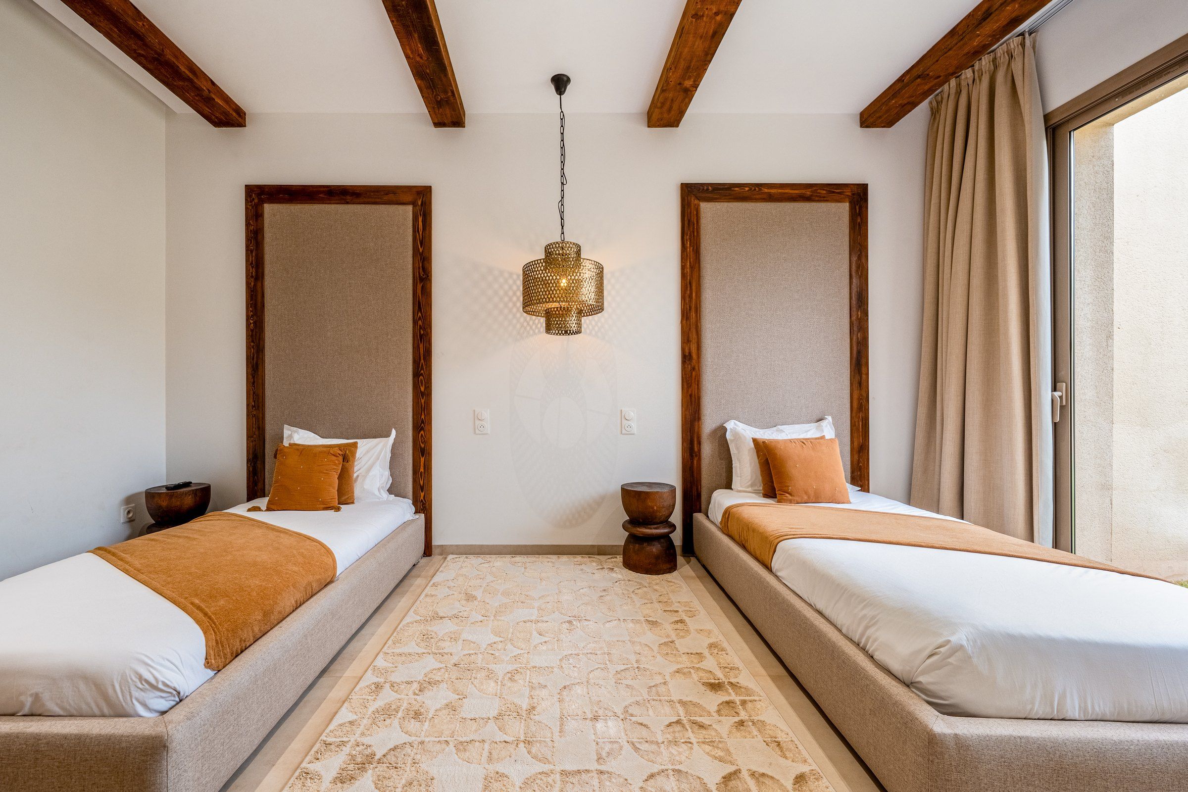A hotel room with two beds, a patterned rug, a hanging light fixture, and wooden beams on the ceiling.