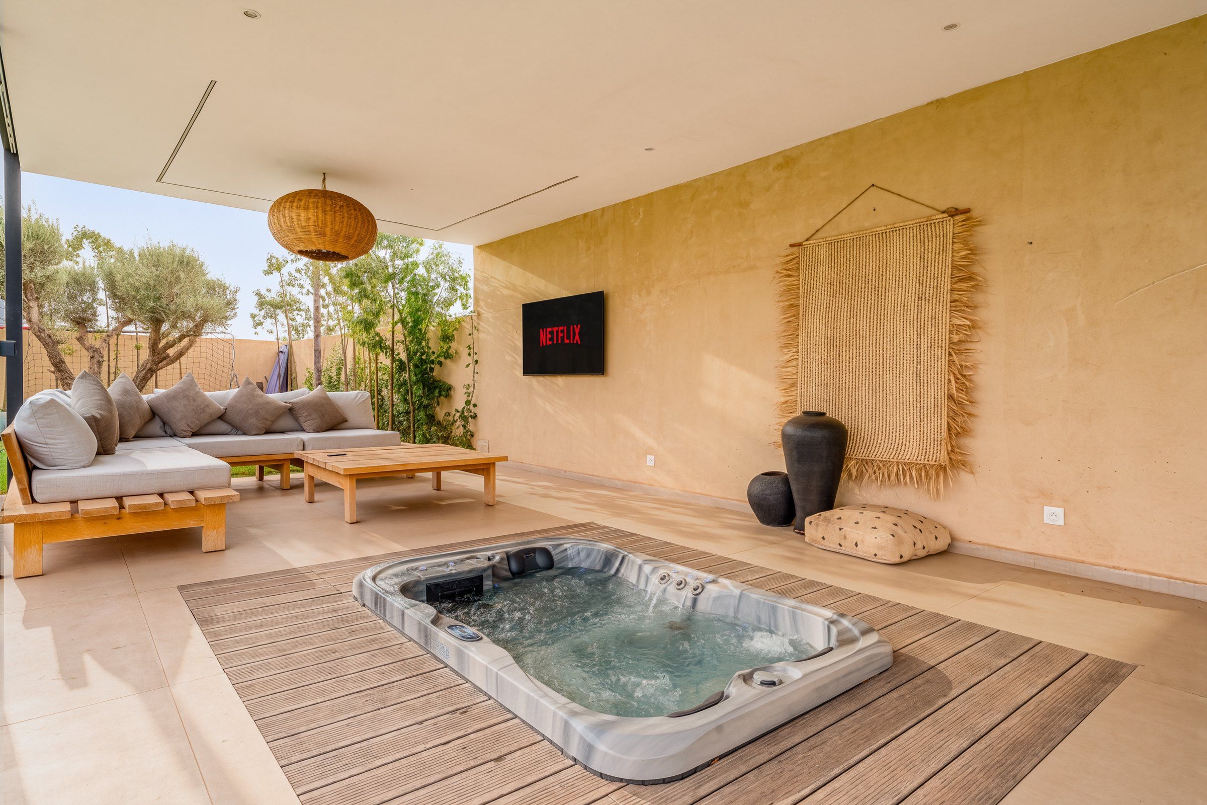 Un espace de vie extérieur moderne comprenant un canapé, une table en bois, un jacuzzi et un téléviseur fixé au mur.
