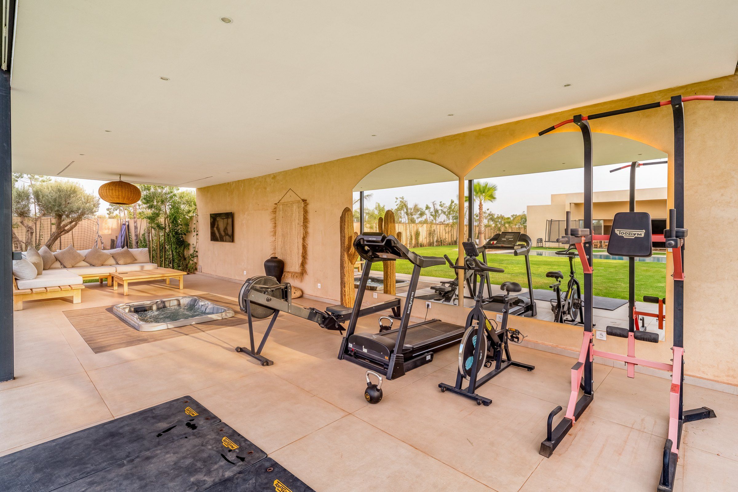 A covered outdoor gym area with exercise equipment, a hot tub, and a seating area with a sofa and table.