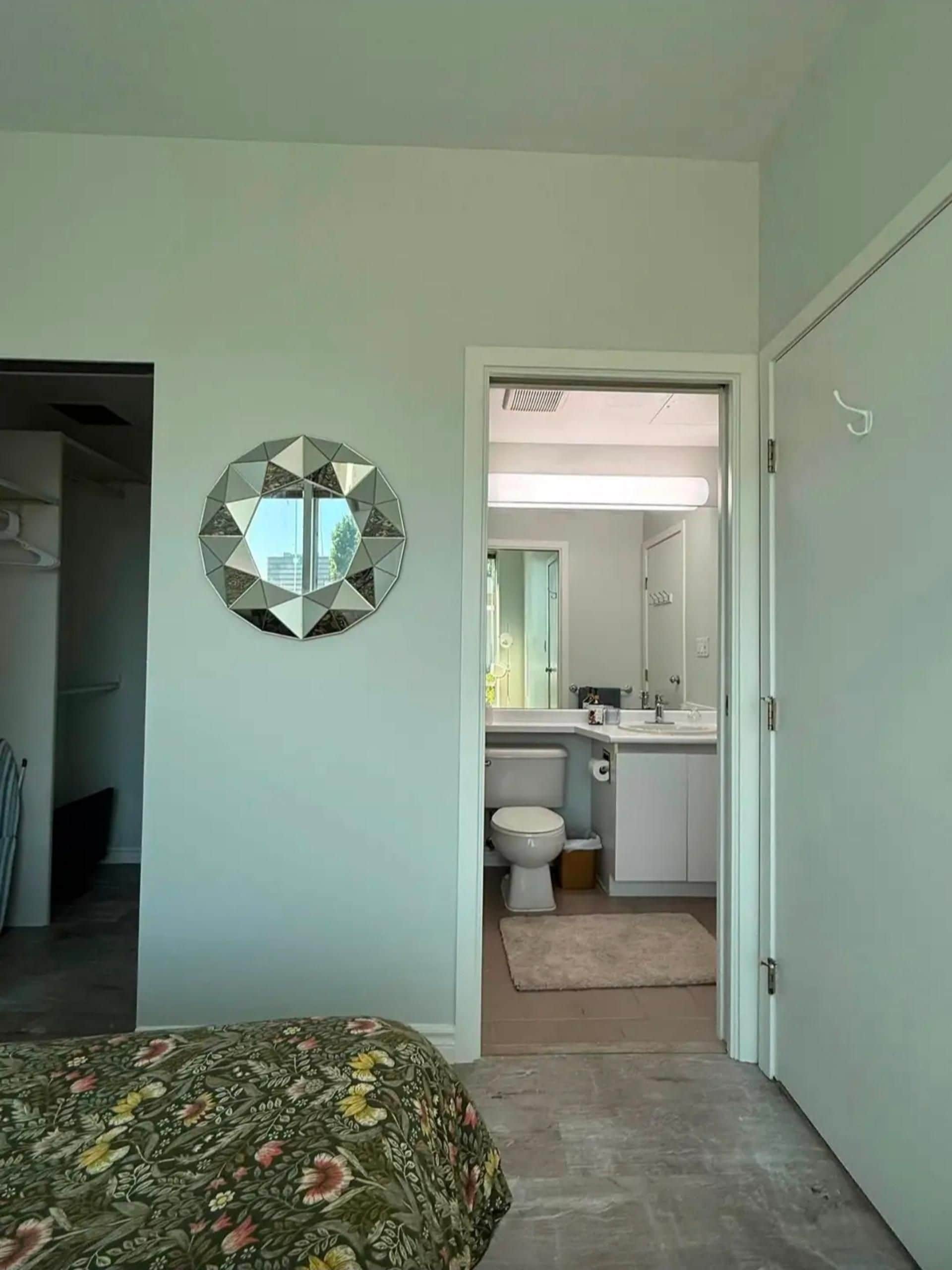 Une chambre à coucher avec un lit, un miroir mural aux motifs géométriques et une vue sur une salle de bain équipée de toilettes et d’un lavabo.