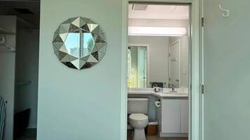 Une chambre à coucher avec un lit, un miroir mural aux motifs géométriques et une vue sur une salle de bain équipée de toilettes et d’un lavabo.