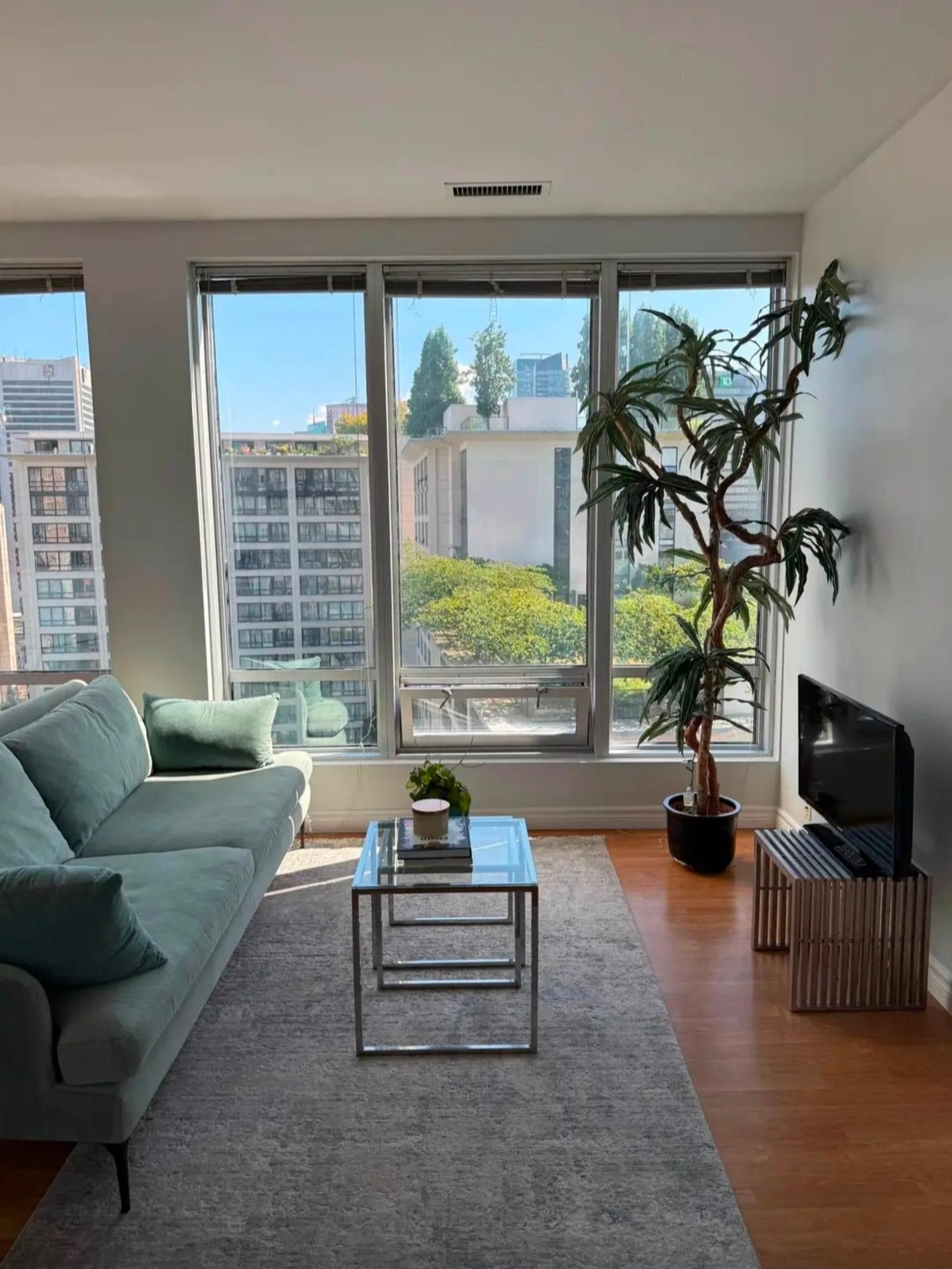 Uma sala de estar moderna com sofá, mesa de centro, rack para TV e uma grande janela com vista para edifícios e árvores.