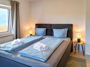 Ein Doppelbett mit zwei Kissen, ein Nachttisch mit Lampe sowie ein Fenster mit Vorhängen.