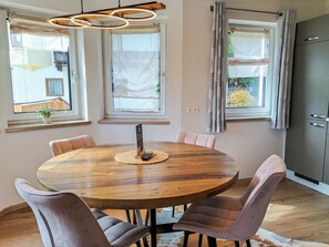 Une table de salle à manger ronde en bois, accompagnée de six chaises, un luminaire suspendu et des fenêtres avec des rideaux.