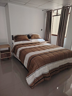 A bedroom with a bed, a nightstand, a window with curtains, and a wall-mounted light.