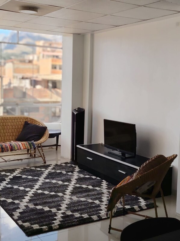 A modern living room with a flat-screen TV on a black stand, a wicker sofa with a colorful throw blanket, and a patterned rug.