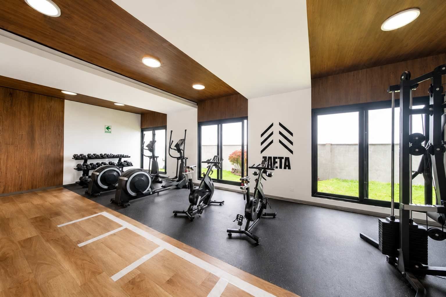 Un gimnasio con equipo de ejercicio, piso de madera y amplias ventanas.