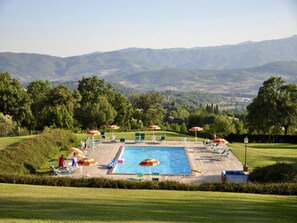 Ein Schwimmbecken mit Liegestühlen und Sonnenschirmen, umgeben von einer gepflegten Rasenfläche und Bäumen, mit Blick auf ferne Hügel.