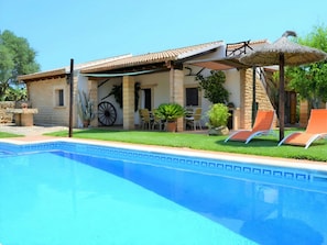 Ein Poolbereich mit klarem, blauem Pool, einer überdachten Terrasse mit Sitzgelegenheiten und einem Steinhaus mit einem Ziegeldach.