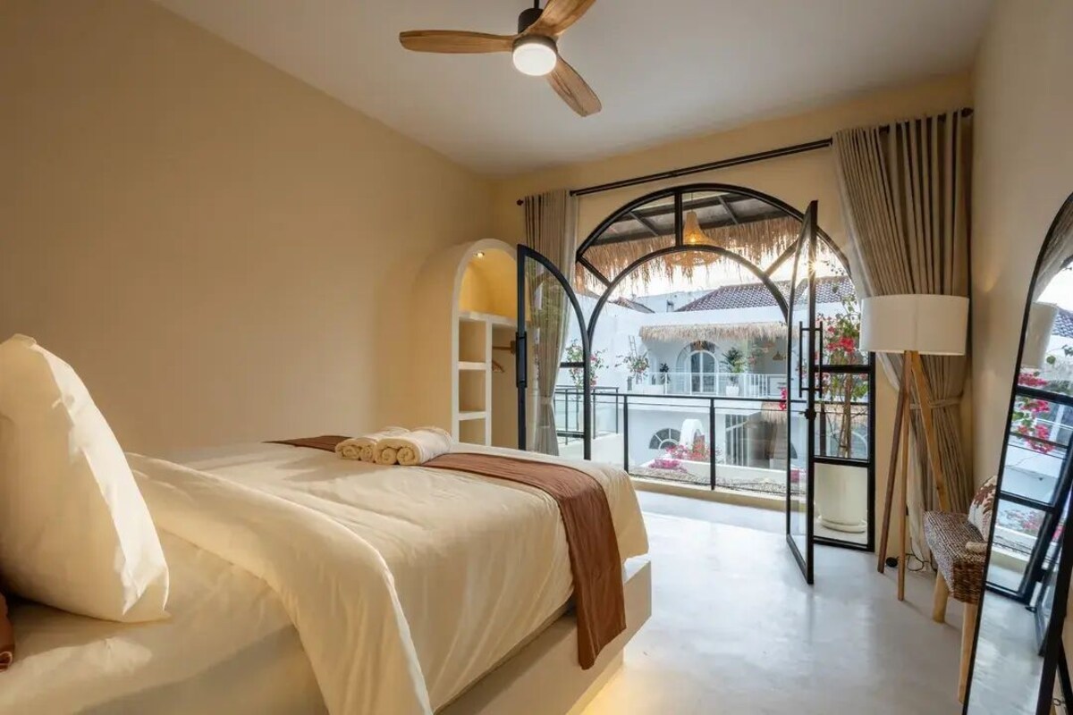 A bedroom with a bed, a ceiling fan, a window with a view of a balcony, and a lamp.