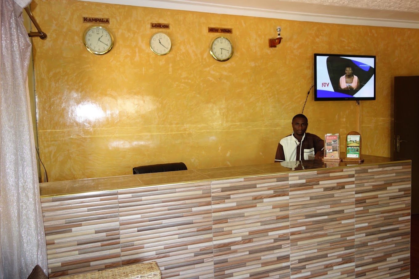 A reception desk with clocks labeled 'Kampala,' 'London,' and 'New York,' and a television screen displaying a news program.