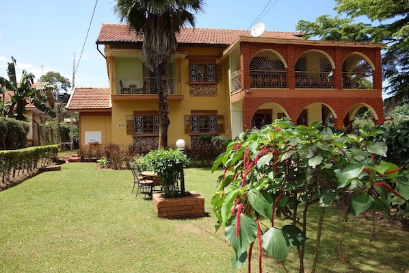 A two-story building with a balcony, a small garden with a table and chairs, and a large plant with red flowers.