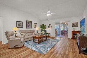 A living room with a sofa, armchair, coffee table, and a fireplace. There is a ceiling fan, a large window, and a door leading to a patio. - Margarita Retreat at Village of Santo Domingo (The Villages)