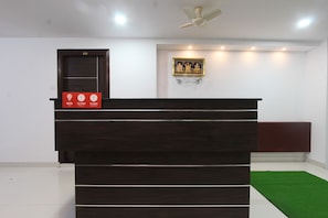 Un mostrador de recepción con una zona de registro, un artefacto religioso enmarcado en la pared y un ventilador de techo.