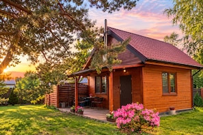 A wooden cabin with a sloped roof, a porch with outdoor seating, and a garden with pink flowers. - Holiday Home for 4 People, Dziwnow (Dziwnów)