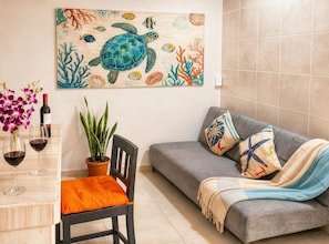 Ein Zimmer mit einem grauen Sofa, einem Holzstuhl mit orangefarbenem Kissen, einem Tisch mit Weingläsern und einer Flasche, einer Topfpflanze und einem Bild von einer Schildkröte und Fischen an der Wand.
