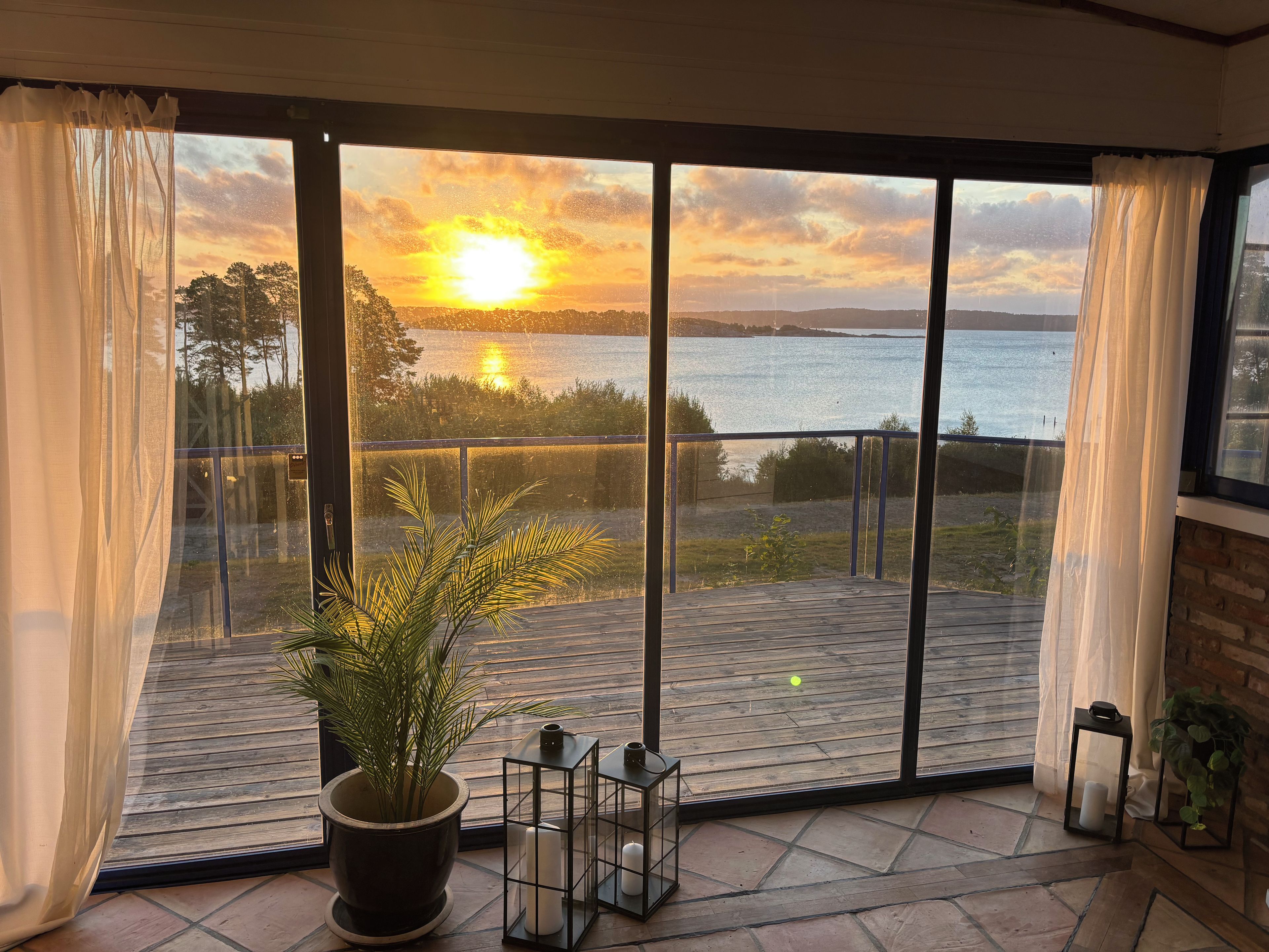 A room with a large window overlooking a body of water and a sunset. There is a potted plant and a small table with a candle on the balcony.