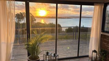 A room with a large window overlooking a body of water and a sunset. There is a potted plant and a small table with a candle on the balcony.