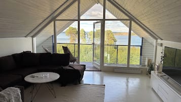 A modern interior with a high ceiling, a dark-colored sofa, a small round table, and a large window offering a view of a forest and water.