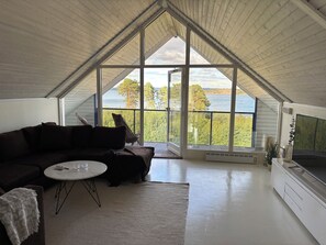 A modern interior with a high ceiling, a dark-colored sofa, a small round table, and a large window offering a view of a forest and water.