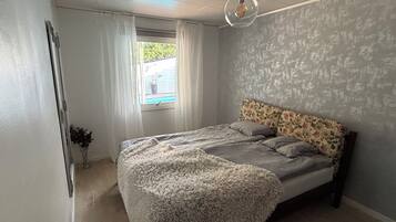 A bedroom with a bed, a window with curtains, a hanging light fixture, and a view of the outdoors.