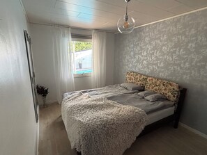 A bedroom with a bed, a window with curtains, a hanging light fixture, and a view of the outdoors.