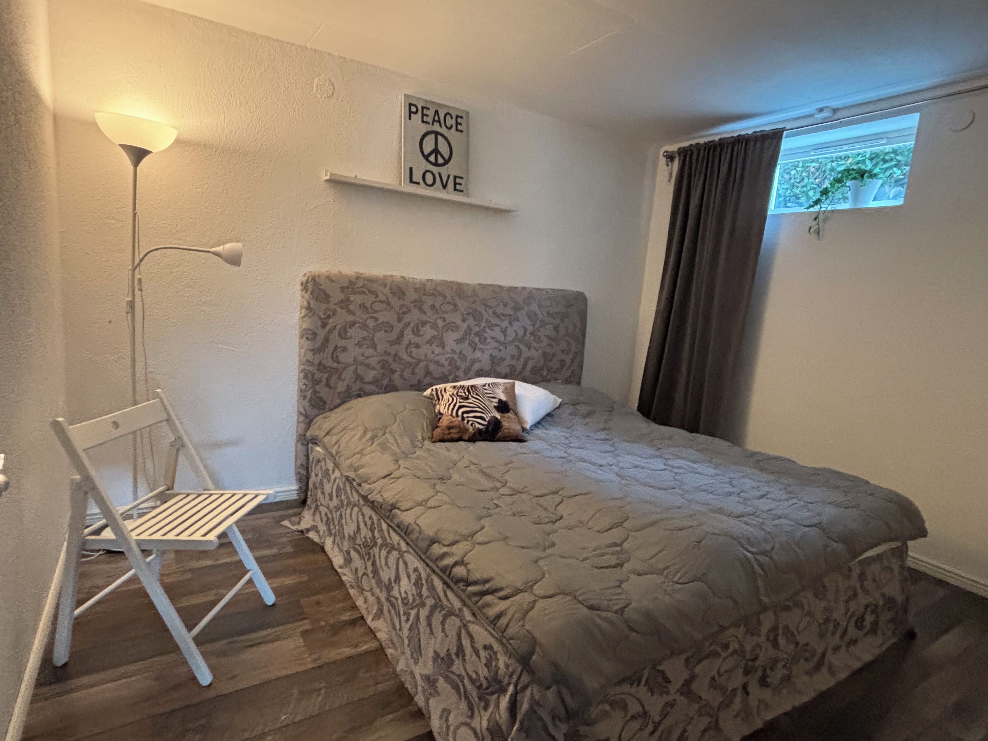 Een slaapkamer met een bed, een stoel, een lamp, een raam met gordijnen, en een 'Peace Love' bord aan de muur.