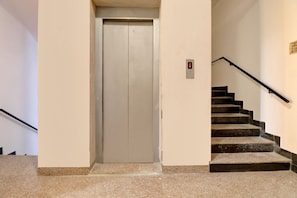 Puerta de ascensor cerrada, escalera con barandilla y panel de alarmas contra incendios en la pared.