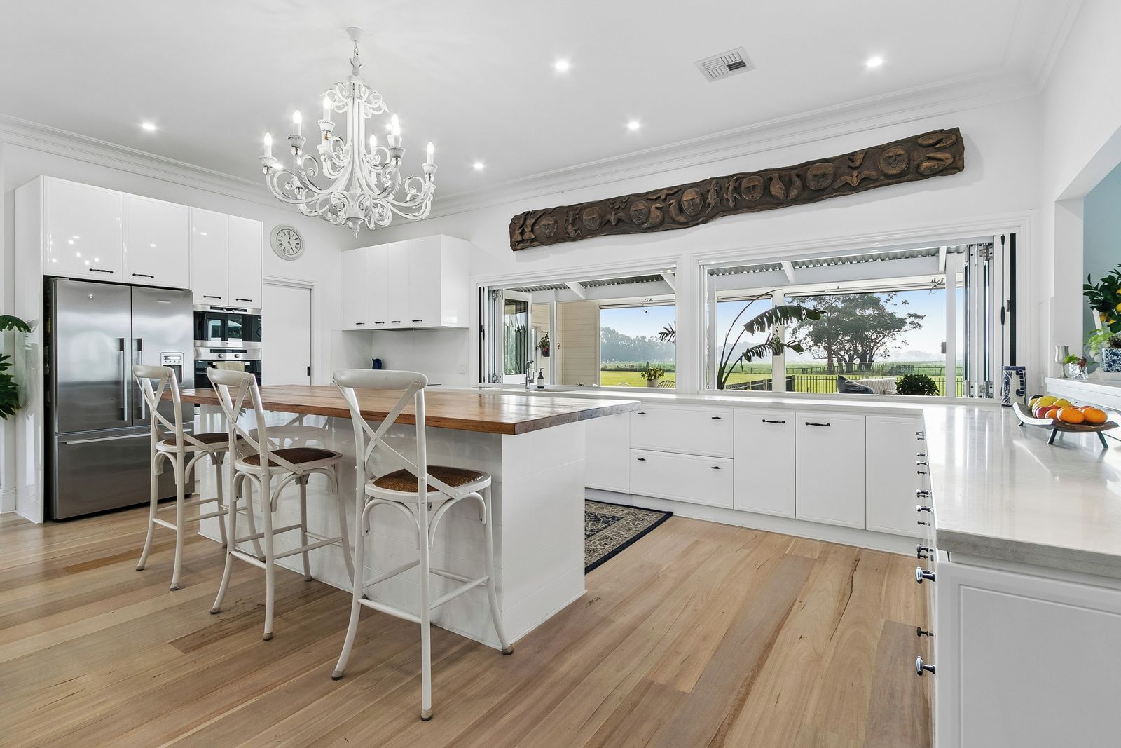 A modern kitchen with white cabinetry, a central island with bar stools, a stainless steel refrigerator, and a view of the outdoors through large windows.