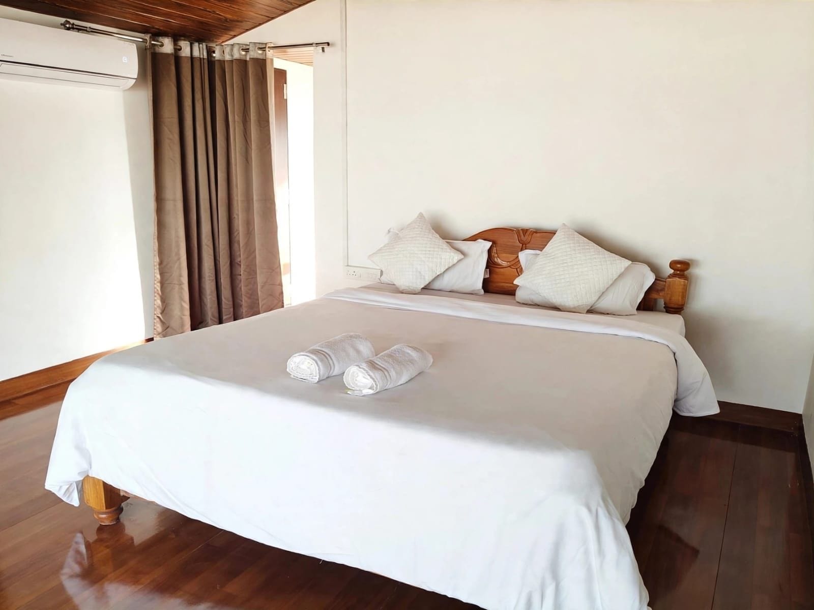 A hotel room with a bed, wooden headboard, white bedding, and a pair of rolled towels on the bed.