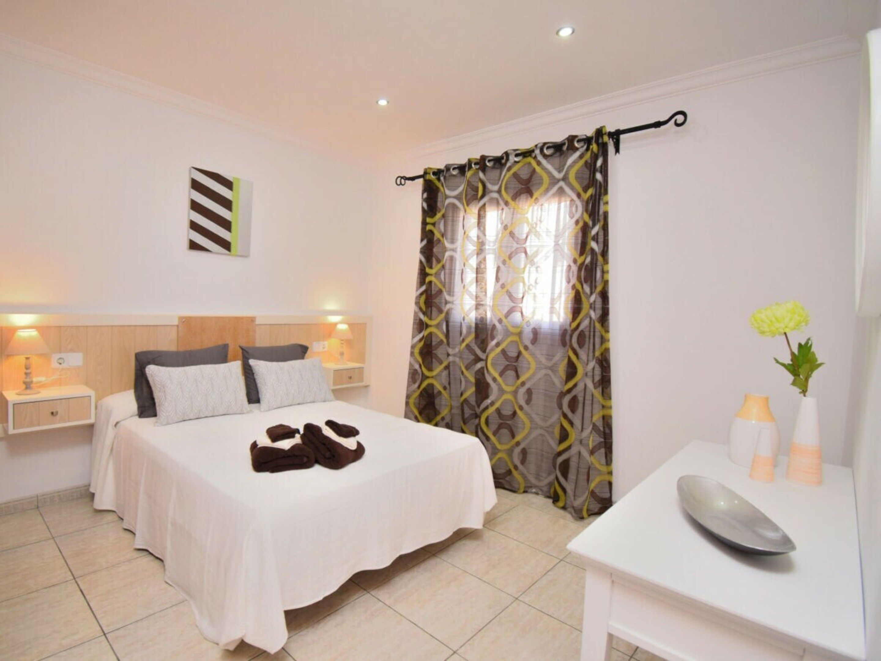 A hotel room with a bed, bedside tables, a desk with a vase, and a curtain.