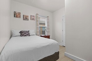 A bedroom with a bed, a nightstand, a lamp, and a window with blinds. - Cozy 3BR Detroit Home | Sleeps 8 | Self Check-in (Detroit)