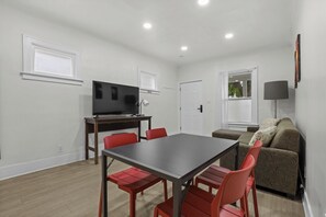 A compact living space with a dining table, red chairs, a sofa, a television, and a window. - Cozy 3BR Detroit Home | Sleeps 8 | Self Check-in (Detroit)