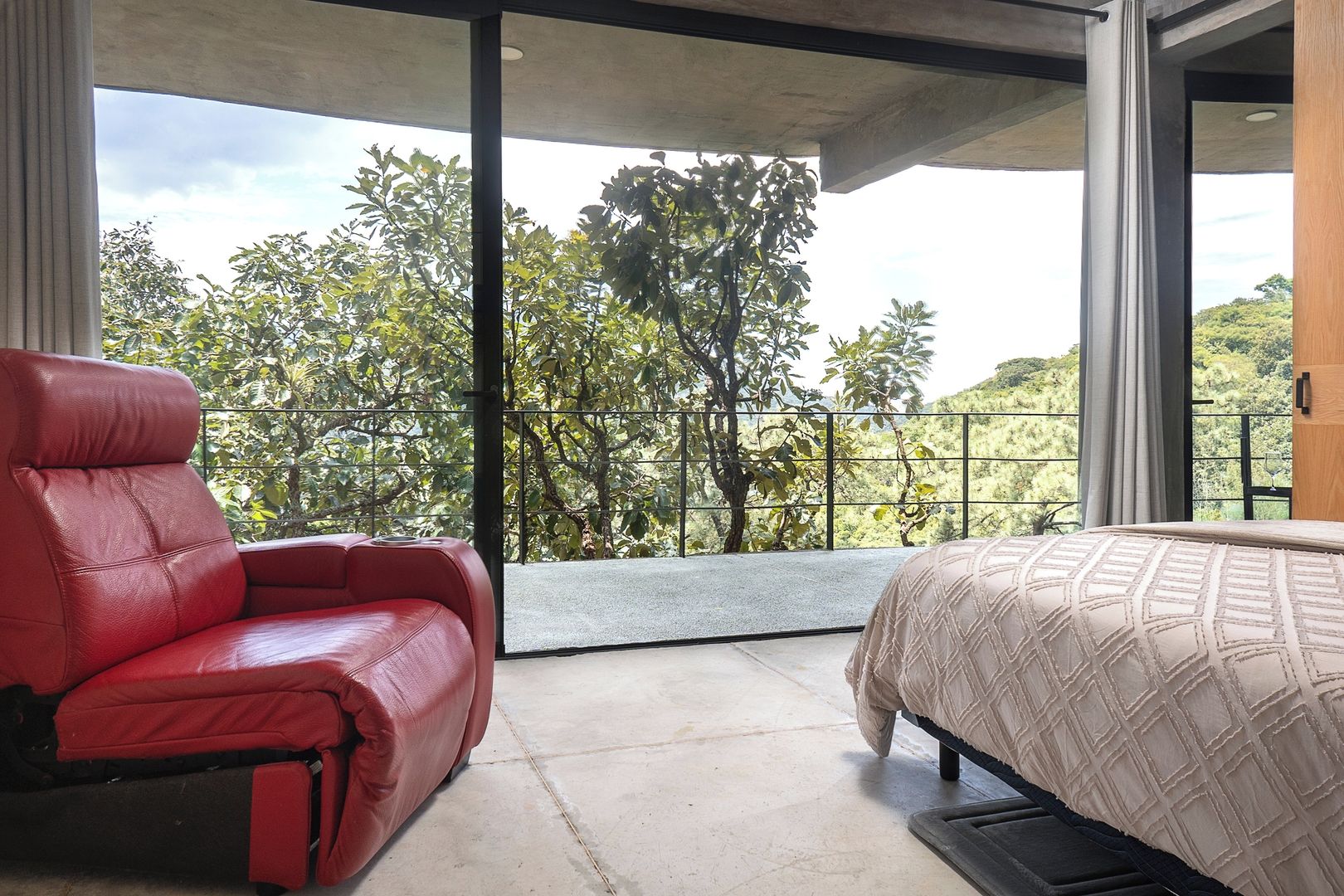 Ein modernes Schlafzimmer mit großem Fenster, einem roten Ledersessel und einem Bett mit gemustertem Überwurf.