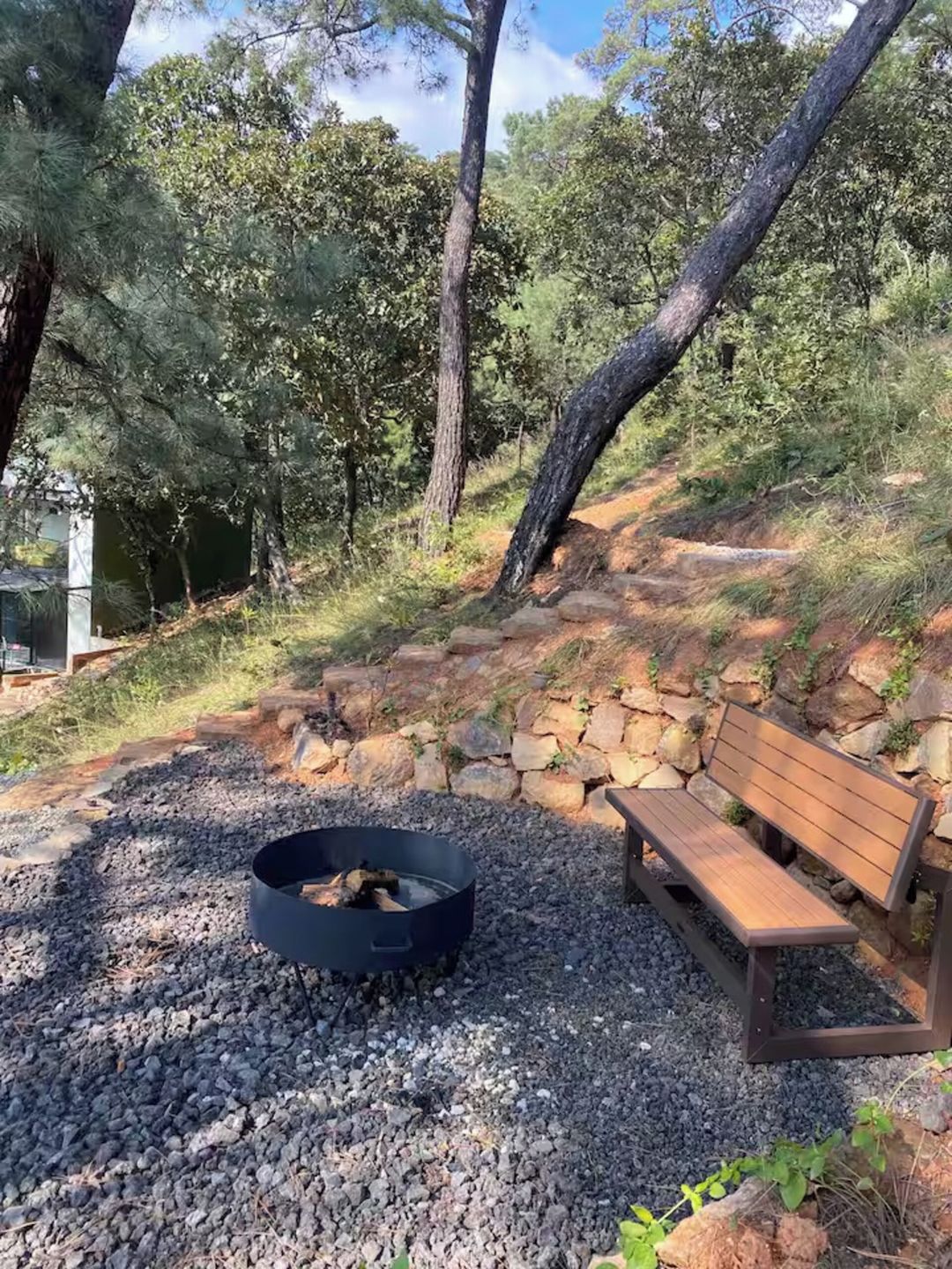 A fire pit with logs, surrounded by gravel, next to a wooden bench and a stone wall.