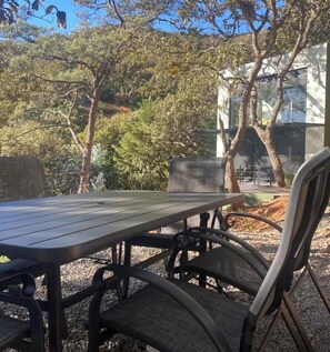 An outdoor dining area with a table and chairs, surrounded by trees and a modern building in the background. - Spectacular Bosque Condo – Lake View – Jacuzzi (Valle de Bravo)