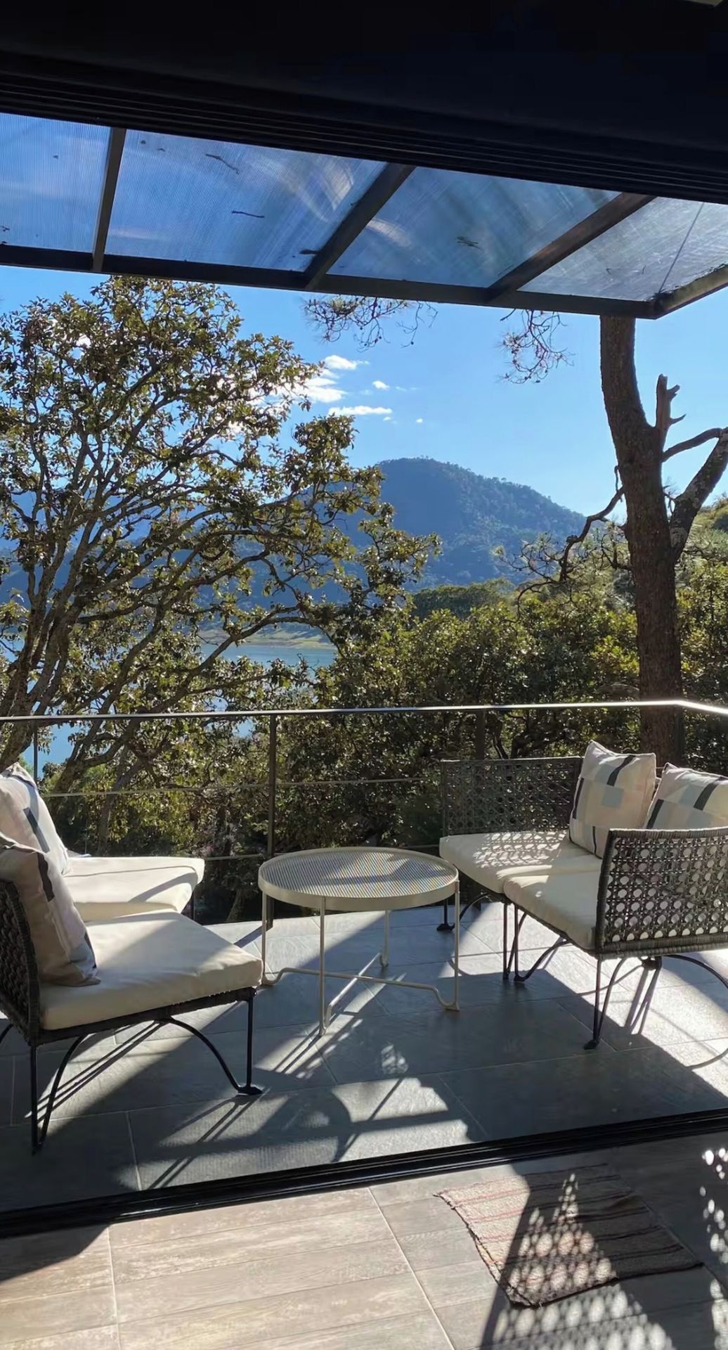 An outdoor patio with a glass ceiling, two lounge chairs, a small table, and a view of a mountainous landscape.