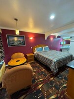 Quarto de hotel com uma cama, uma cadeira, uma mesinha, televisão fixada na parede e um quadro na parede.