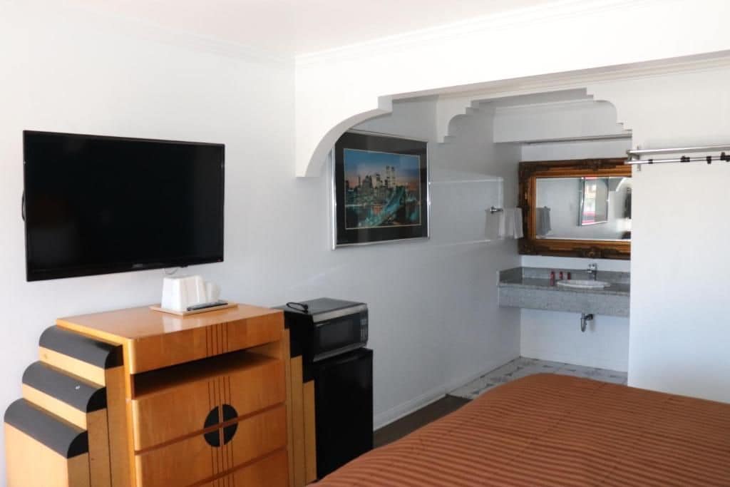A hotel room with a flat-screen TV, a wooden dresser, a small trash bin, a bed with a striped bedspread, a bathroom with a sink and mirror, and a cityscape picture on the wall.
