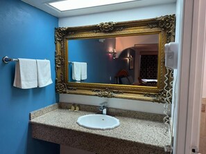 Een badkamer met een grote, versierde spiegel, een granieten aanrecht, een witte wastafel en een handdoekhouder met twee handdoeken.